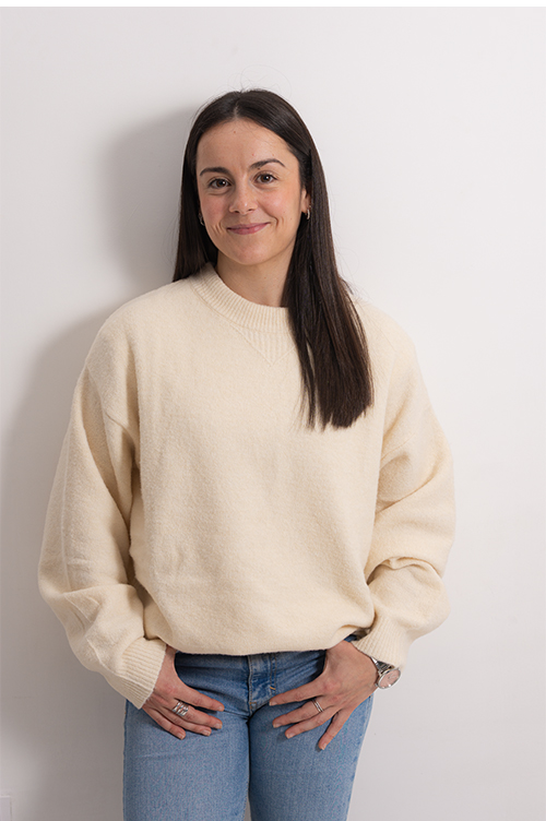 Ana Perez, gimnasta. Mujer de piel clara posando frente a una pared lisa de color gris claro. Tiene el cabello largo, lacio y oscuro, con raya al medio, y una leve sonrisa.  Viste un suéter de punto color crema/beige con cuello redondo y puños acanalados, y jeans azules. Lleva un reloj metálico en una muñeca y anillos en los dedos. La postura es de frente, con las manos apoyadas cerca de la cintura. No hay texto visible en la imagen.