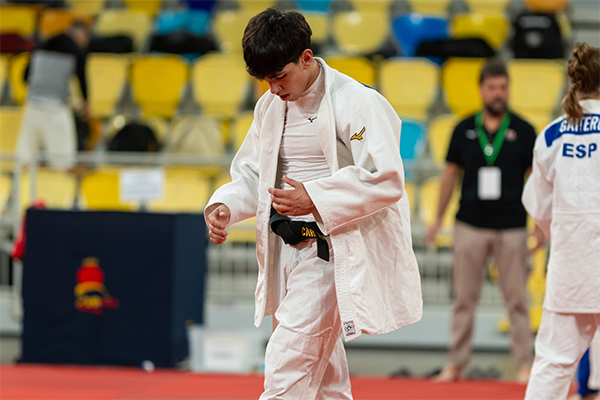 Carlos martin Un judoca joven con kimono blanco en un tatami de competición. Lleva debajo una camiseta ajustada blanca y un cinturón negro que está sujetando o ajustando con las manos. Tiene el cabello corto y oscuro, la cabeza ligeramente inclinada hacia abajo. En la parte derecha, de espaldas, hay otra persona con kimono blanco con las letras “ESP” en azul en la espalda. Al fondo se ven gradas con asientos amarillos y algunas personas de pie, una de ellas con acreditación colgada al cuello.