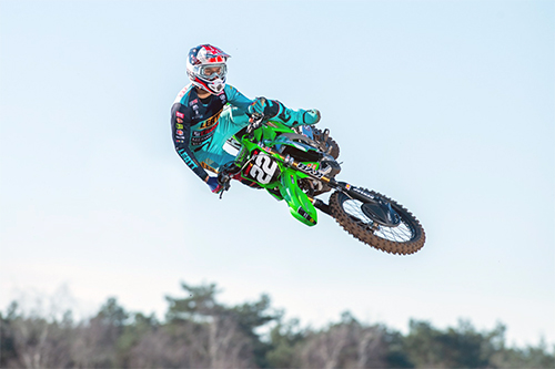 La imagen muestra al motociclista David Braceras realizando un salto en el aire con una motocicleta de motocross. El piloto lleva un traje de color turquesa y un casco con diseño en rojo, blanco y azul. La motocicleta es de color verde y tiene el número 29 visible en la parte delantera. El fondo es un cielo claro con algunos árboles desenfocados en la parte inferior de la imagen.