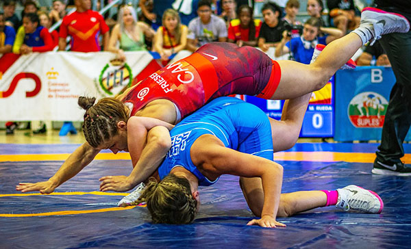 Lucha libre femenina. En la imagen, dos luchadoras están compitiendo en un torneo de lucha libre en un gimnasio. Una de ellas, vestida con un leotardo rojo, está encima de la otra, que lleva un leotardo azul. La luchadora roja sostiene a su oponente, intentando realizar un movimiento para tumbarla y dominarla en el suelo. Ambas parecen estar empleando mucha fuerza y destreza física. Al fondo, se ve público observando el combate y varias personas emocionadas, junto con pancartas y carteles en las paredes. La escena transmite mucha energía y acción deportiva.