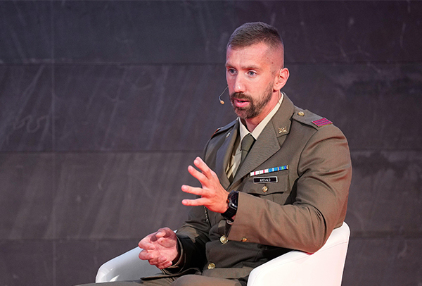 La imagen muestra a Carlos Arévalo, piragüista olímpico, vestido con un uniforme militar mientras está sentado en una silla blanca. El uniforme es de color verde oliva y tiene varias insignias y medallas en el pecho. Lleva una corbata de color claro y tiene un micrófono de diadema en su oreja derecha. Carlos Arévalo tiene el cabello corto y barba. Está gesticulando con la mano derecha mientras habla. El fondo es de color oscuro, posiblemente una pared o un telón.