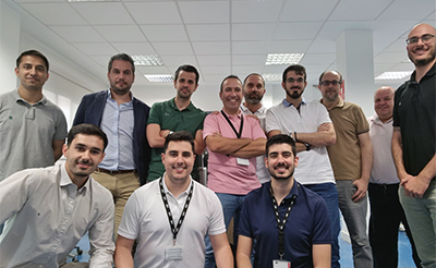 La imagen muestra a un grupo de 12 hombres posando para una foto en lo que parece ser una oficina. Están de pie y sentados, y todos están sonriendo. La mayoría de ellos lleva ropa casual, como camisas de manga corta y pantalones. El fondo muestra un techo con paneles de luz y paredes blancas, lo que sugiere un ambiente de trabajo moderno y bien iluminado.