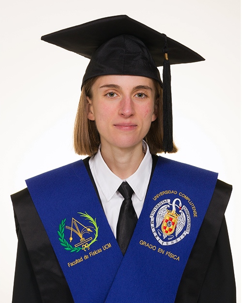 La imagen muestra a una persona joven vestida con una toga y birrete de graduación. Lleva una banda azul sobre los hombros. En la banda, a la izquierda, se puede ver el logo de la Facultad de Físicas de la Universidad Complutense de Madrid (UCM), y a la derecha, el escudo de la universidad con las palabras "Grado en Física". La persona tiene el cabello corto y lacio, y está mirando directamente a la cámara con una expresión neutral.