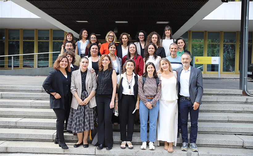 Grupo de personas posando en las escaleras de entrada de un edificio moderno. La mayoría son mujeres; hay dos hombres en la esquina derecha de la imagen. Todas las personas están vestidas de manera formal o semiformal, algunas con trajes y otras con ropa más casual. Están sonriendo y mirando a la cámara, dando la impresión de ser una foto grupal de un evento, reunión o curso. Al fondo, se ve una fachada con ventanas grandes y un letrero amarillo con logotipo y texto.