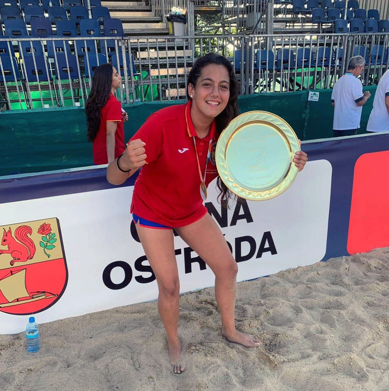 La imagen muestra a Carolina Jiménez, en una playa, posando con una gran bandeja dorada que parece ser un trofeo. Está vestida con una camiseta roja y pantalones cortos azules. Lleva una medalla colgada al cuello y está descalza sobre la arena. La joven tiene el puño derecho levantado en señal de victoria y una gran sonrisa en su rostro. Detrás de ella, hay una tribuna con asientos vacíos y algunas personas de pie. También se puede ver una botella de agua en la arena cerca de ella. En la barrera que la separa de la tribuna, hay un escudo con un dibujo de una ardilla y una rosa.