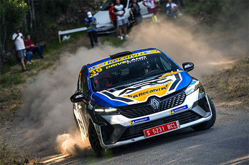 Coche de rally Renault azul y amarillo, con matrícula roja, derrapando en una curva de asfalto mientras levanta polvo. Hay espectadores al fondo, algunos de pie y otros sentados, observando la acción desde el borde del camino y entre los árboles. El coche tiene pegatinas de patrocinadores y el número 39 en el parabrisas. El piloto es José Javier Pérez Briones. La escena transmite velocidad y emoción en una competencia automovilística.