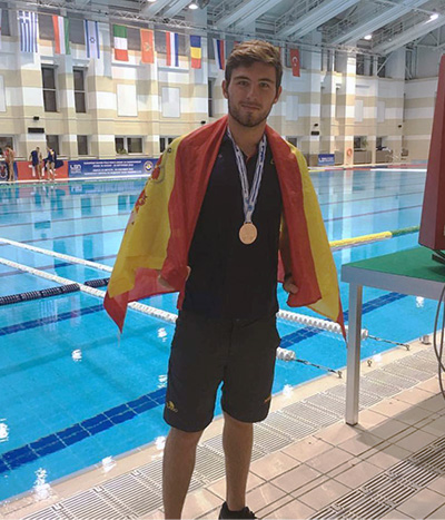 La imagen muestra a José María Tenorio, jugador de waterpolo, de pie junto a una piscina cubierta. Está envuelto en una bandera de España y lleva una medalla de bronce colgada al cuello. La piscina tiene varias líneas de carriles y el fondo es azul. En la parte superior de la imagen, se pueden ver varias banderas de diferentes países colgadas en la pared. El joven parece estar en una competencia de natación, dado el entorno y la medalla que lleva.