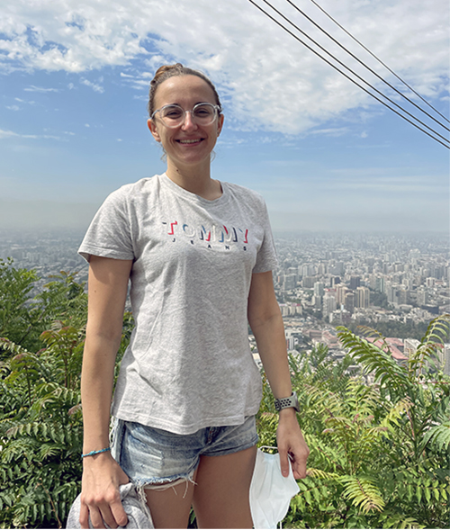 La imagen muestra a María Delgado, nadadora paralímpica, una mujer joven de pie al aire libre con una ciudad en el fondo. Ella lleva gafas, una camiseta gris con el logo de "Tommy Jeans" y pantalones cortos de mezclilla. Tiene el cabello recogido en una coleta y está sonriendo. En su muñeca izquierda lleva una pulsera azul y un reloj, y en su mano derecha sostiene una mascarilla blanca. Detrás de ella hay vegetación y se puede ver una vista panorámica de la ciudad con muchos edificios y un cielo parcialmente nublado.