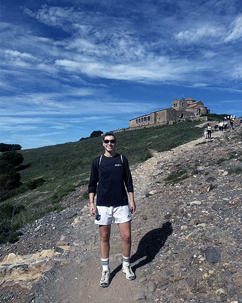 En la imagen aparece una persona de pie en un sendero de montaña, bajo un cielo azul con algunas nubes. Está sonriente, lleva gafas de sol, una camiseta de manga larga negra y pantalones cortos blancos, con calcetas blancas y zapatillas deportivas. Se encuentra en primer plano y parece estar disfrutando del paseo. Al fondo, sobre la colina, se observa un edificio de piedra con tejado inclinado, que parece ser una iglesia o un monasterio. También se ven varias personas más caminando por el sendero, cerca del edificio. La vegetación alrededor es baja y el terreno es pedregoso. Es la taekwondista Tania Castiñeira.