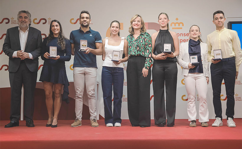 premiados carrera dual 2025 : Un grupo de ocho personas de pie sobre un escenario alfombrado de rojo. Siete de ellas sostienen medallas o premios en sus manos y sonríen hacia la cámara. Todos están vestidos de manera formal o semiformal, con ropa elegante. El fondo muestra un mural con logotipos y textos, incluyendo un símbolo de corona, letras “CSD” y el texto “Consejo Superior de Deportes”. Parece una ceremonia de premiación o reconocimiento deportivo.