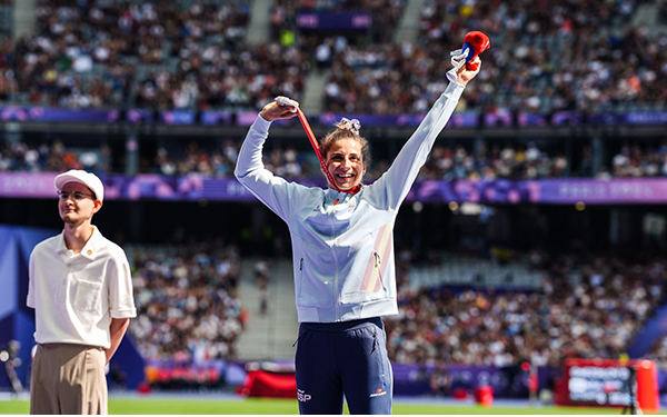 La imagen muestra a Sara Martínez, la atleta paralímpica, en pleno salto durante una competencia de atletismo en un estadio lleno de espectadores. La atleta lleva un uniforme rojo y blanco con el nombre "Martínez" visible en su camiseta. Está en el aire con los brazos levantados y las piernas dobladas, en lo que parece ser un salto de longitud. En el fondo, se puede ver una pista de atletismo y una pantalla grande que muestra información del evento. El estadio está lleno de personas, y hay un juez o entrenador sentado a un lado observando el salto.