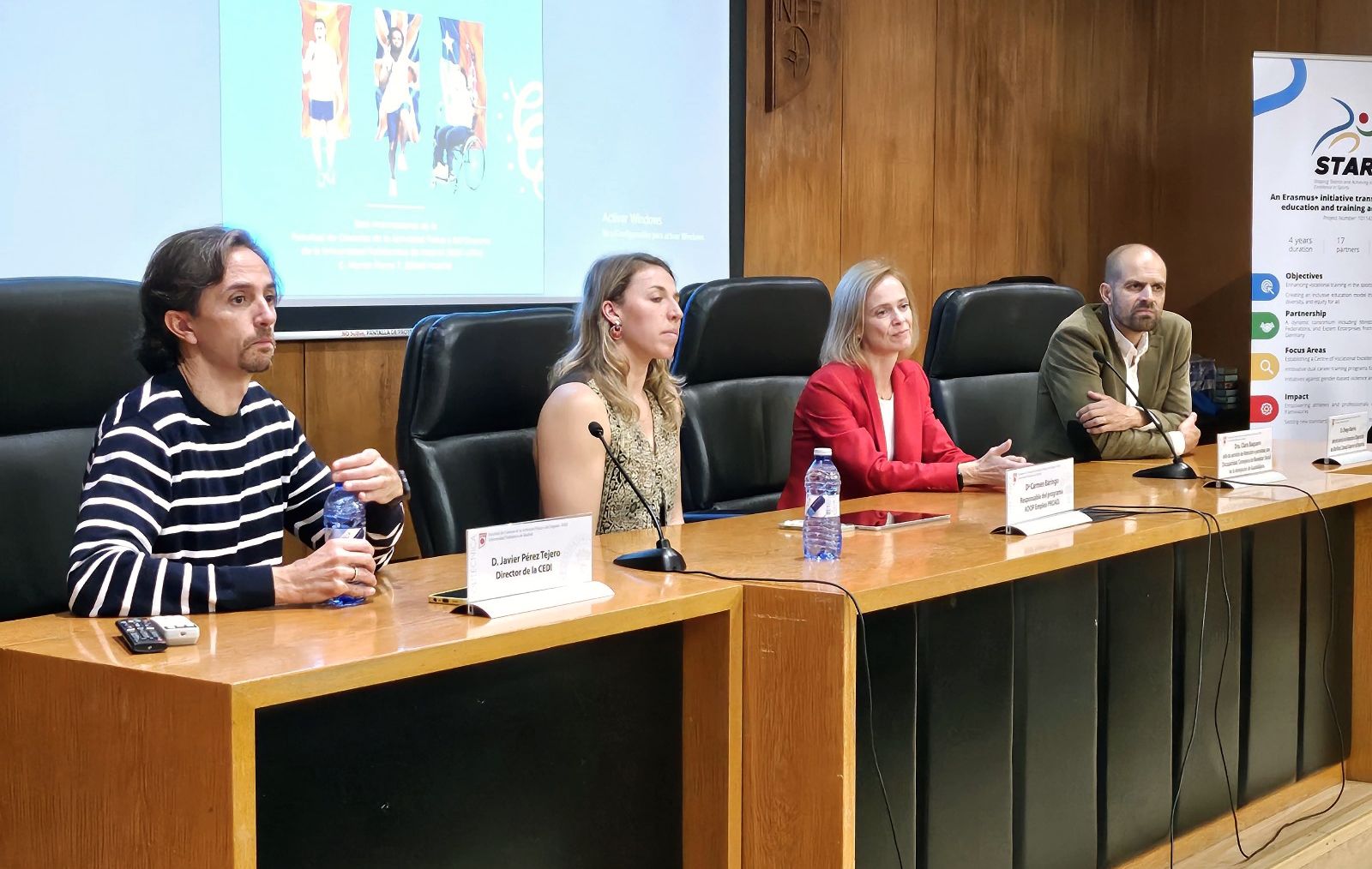 La imagen muestra a cuatro personas sentadas en una mesa de conferencias en un auditorio. De izquierda a derecha, el primer hombre lleva una camiseta de rayas horizontales blancas y negras y sostiene una botella de agua. Frente a él hay un cartel que dice "D. Javier Pérez Tejero, Director de la CEDI". La segunda persona es una mujer con un vestido sin mangas de color claro, y también tiene un micrófono frente a ella. La tercera persona es una mujer con una chaqueta roja, y frente a ella hay una botella de agua y un dispositivo electrónico. La cuarta persona es un hombre con una chaqueta marrón y camisa blanca. En la pared detrás de ellos hay una pantalla con imágenes y texto, y a la derecha hay un cartel vertical con información sobre un proyecto Erasmus llamado "STAR".