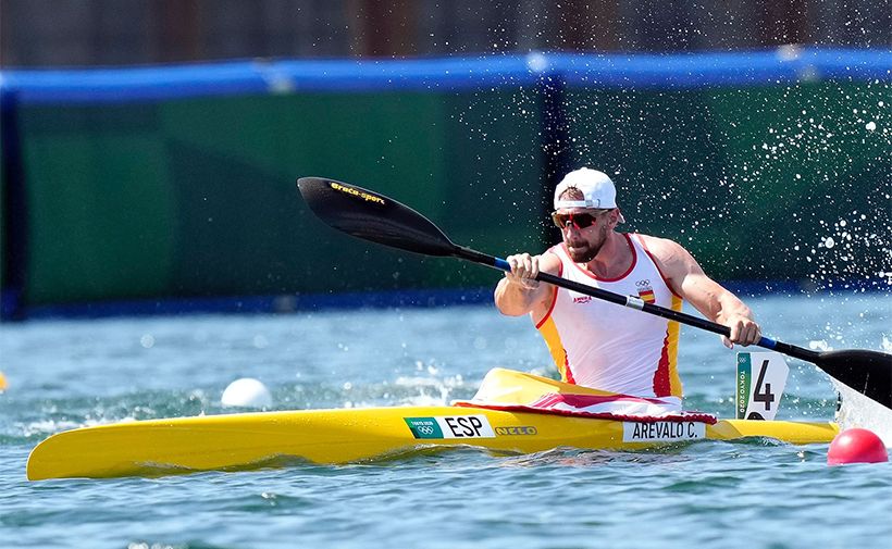 La imagen muestra al deportista Carlos Arévalo participando en una competencia de piragüismo. Está remando en una piragua amarilla en el agua. Lleva una camiseta sin mangas blanca con detalles en rojo y amarillo, que son los colores de España, y un gorro blanco. En la piragua se puede ver la inscripción "ESP" y "AREVALO C." junto al número 4. Carlos Arévalo está concentrado en su esfuerzo, y se pueden ver salpicaduras de agua alrededor debido a la velocidad y fuerza con la que está remando.