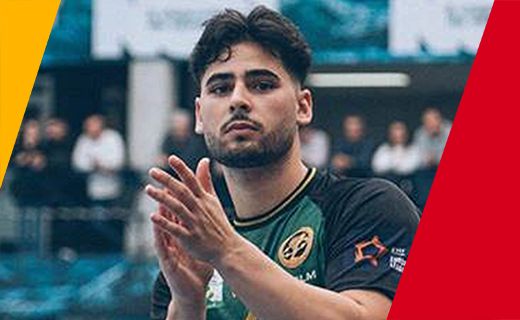 La imagen muestra a Arnau Fernández, jugador de balonmano,  es un joven con cabello oscuro y barba ligera, aplaudiendo. Lleva puesta una camiseta deportiva de color negro con detalles en verde y un logotipo dorado en el pecho. En el fondo, se puede ver a varias personas desenfocadas, lo que sugiere que está en un evento o lugar público. A los lados de la imagen hay franjas de color amarillo y rojo.