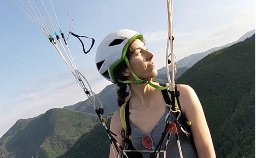 La imagen muestra a Azahara Velasco, una deportista de parapente, mientras practica este deporte. Lleva puesto un casco blanco con detalles verdes y un arnés de seguridad. Está mirando hacia su derecha, con una expresión serena en su rostro. El fondo muestra un paisaje montañoso con vegetación verde bajo un cielo despejado. Las cuerdas del parapente son visibles, sujetas al arnés que lleva puesto.