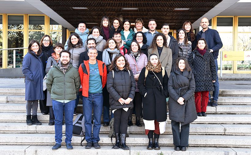 La imagen muestra a un grupo de 25 personas posando en las escaleras frente a un edificio. La mayoría de las personas están abrigadas con chaquetas y bufandas, lo que sugiere que hace frío. Hay una mezcla de hombres y mujeres, y todos están sonriendo. El edificio detrás de ellos tiene grandes ventanas con marcos amarillos y un techo con un diseño de rejilla. Parece ser una foto grupal posiblemente tomada en un contexto académico o profesional.