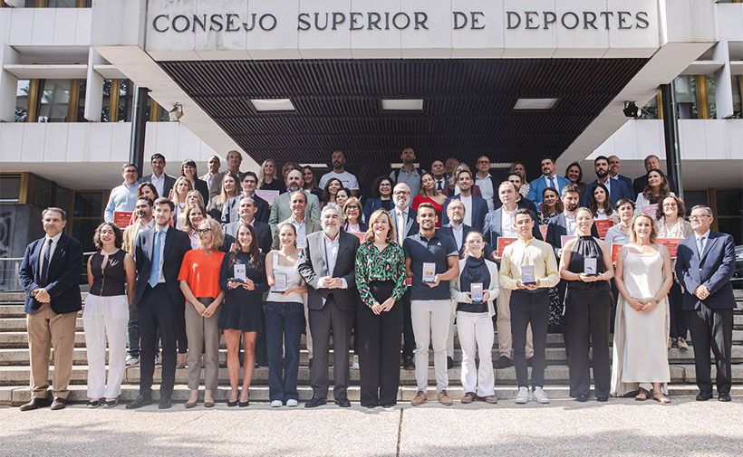 Grupo numeroso de personas posando en las escaleras de un edificio oficial. En la parte superior del edificio, se lee claramente el letrero "CONSEJO SUPERIOR DE DEPORTES". La imagen muestra a personas de diferentes edades, hombres y mujeres, la mayoría de ellos vestidos de manera formal o semiformal. Varias personas sostienen trofeos o placas, lo que sugiere que se trata de una ceremonia de entrega de premios o reconocimientos. La atmósfera es solemne y de celebración. Al fondo, se puede ver la arquitectura moderna del edificio y algunos ventanales.