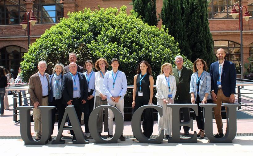 Grupo de personas posando al aire libre frente a un gran arbusto verde y un edificio de ladrillo. Están de pie detrás de grandes letras que forman la palabra "UAO CEU". La mayoría de las personas llevan colgadas credenciales azules, lo que sugiere que podrían estar participando en un evento o conferencia. Están vestidos de manera formal o semiformal, y el clima parece ser soleado.