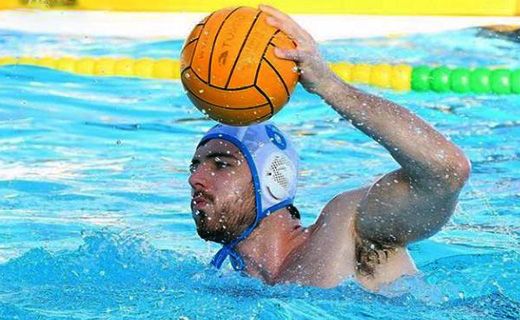 La imagen muestra a José María Tenorio jugador de waterpolo en acción dentro de una piscina. El jugador está parcialmente sumergido en el agua y lleva un gorro de waterpolo azul con el número 3 visible. Está levantando un brazo y sosteniendo una pelota de waterpolo naranja sobre su cabeza, como si estuviera a punto de lanzar la pelota. En el fondo, se pueden ver las líneas de flotadores de colores que delimitan las áreas de juego en la piscina.