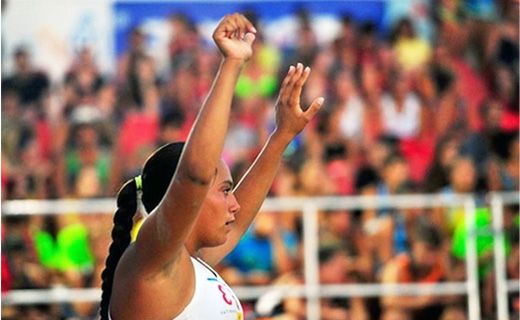 La imagen muestra a Carolina Jiménez, jugadora de balonmano, una mujer con el cabello oscuro recogido en una trenza, levantando ambos brazos en el aire. Ella parece estar en un evento deportivo, posiblemente una competición, ya que lleva una camiseta sin mangas. En el fondo, se puede ver una multitud de personas desenfocadas, lo que sugiere que hay muchos espectadores presentes. Carolina parece estar celebrando o saludando a la multitud.