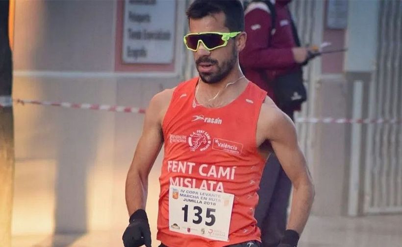 La imagen muestra al atleta Iván Pajuelo participando en una carrera. Lleva una camiseta sin mangas de color naranja con el texto "FENT CAMÍ MISLATA" y "Valencia". También lleva gafas de sol deportivas de color amarillo y negro, y guantes negros. En su camiseta, hay un número de corredor, "135", y debajo se lee "IV COPA LEVANTE MARCHA EN RUTA JUMILLA 2018". Iván Pajuelo parece estar concentrado mientras corre. Al fondo, se puede ver a otra persona vestida con ropa deportiva y una cinta de seguridad que delimita el área de la carrera.