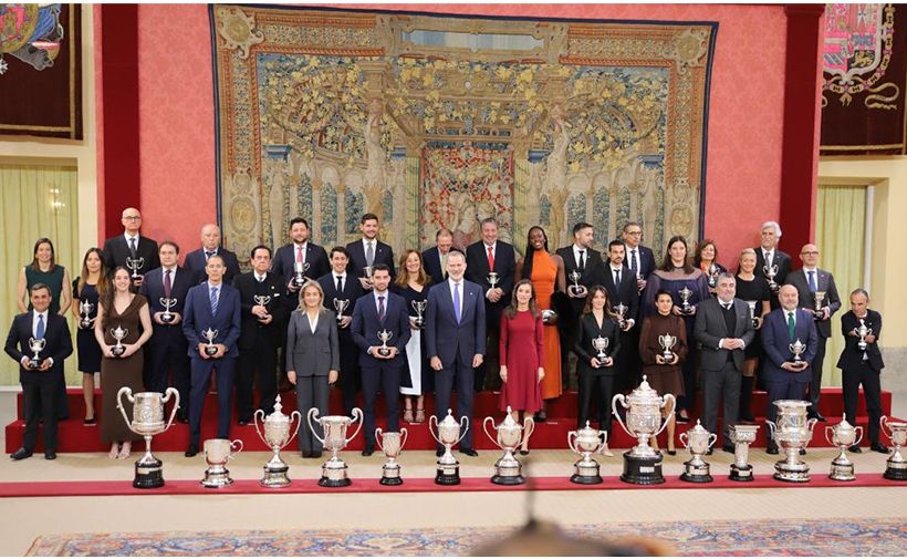 Grupo numeroso de personas posando con trofeos sobre una tarima roja. En total hay unas cuarenta personas organizadas en tres filas: la primera fila está de pie al borde de la tarima, la segunda fila algo elevada y la tercera aún más alta, formando una especie de escalera. La mayoría viste ropa formal: hombres con trajes oscuros y corbatas, y mujeres con vestidos o trajes de chaqueta en colores sobrios, salvo algún vestido de color más vivo como un naranja intenso o un rojo. Casi todos sostienen copas plateadas pequeñas en las manos. Delante de la primera fila, alineados en el suelo, hay muchos trofeos grandes, casi todos de metal plateado brillante, con diferentes formas y tamaños: varias copas con asas grandes, otras más estilizadas y algunas jarras o tazas altas, formando una fila continua. En el centro de la primera fila destaca un hombre alto con traje azul oscuro y corbata azul claro, y a su lado, ligeramente a la derecha, una mujer con vestido rojo de manga larga; ambos están en posición central respecto al grupo. El fondo es una pared decorada con un gran tapiz de estilo clásico que ocupa casi todo el ancho de la imagen. El tapiz tiene muchos detalles, con figuras humanas y elementos arquitectónicos en tonos dorados, verdes y azules. El entorno transmite un ambiente solemne y ceremonial, típico de un acto oficial de entrega de premios.