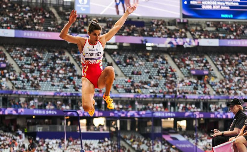 La imagen muestra a Sara Martínez, la atleta paralímpica, en un estadio lleno de espectadores. Sara está sonriendo y levantando los brazos en señal de victoria mientras sostiene una medalla con una cinta roja. Lleva puesta una chaqueta deportiva de color claro y pantalones oscuros. A su lado, hay otra persona con una gorra blanca y una camisa blanca, que parece estar observando la ceremonia. El estadio está lleno de gente y hay una atmósfera de celebración.