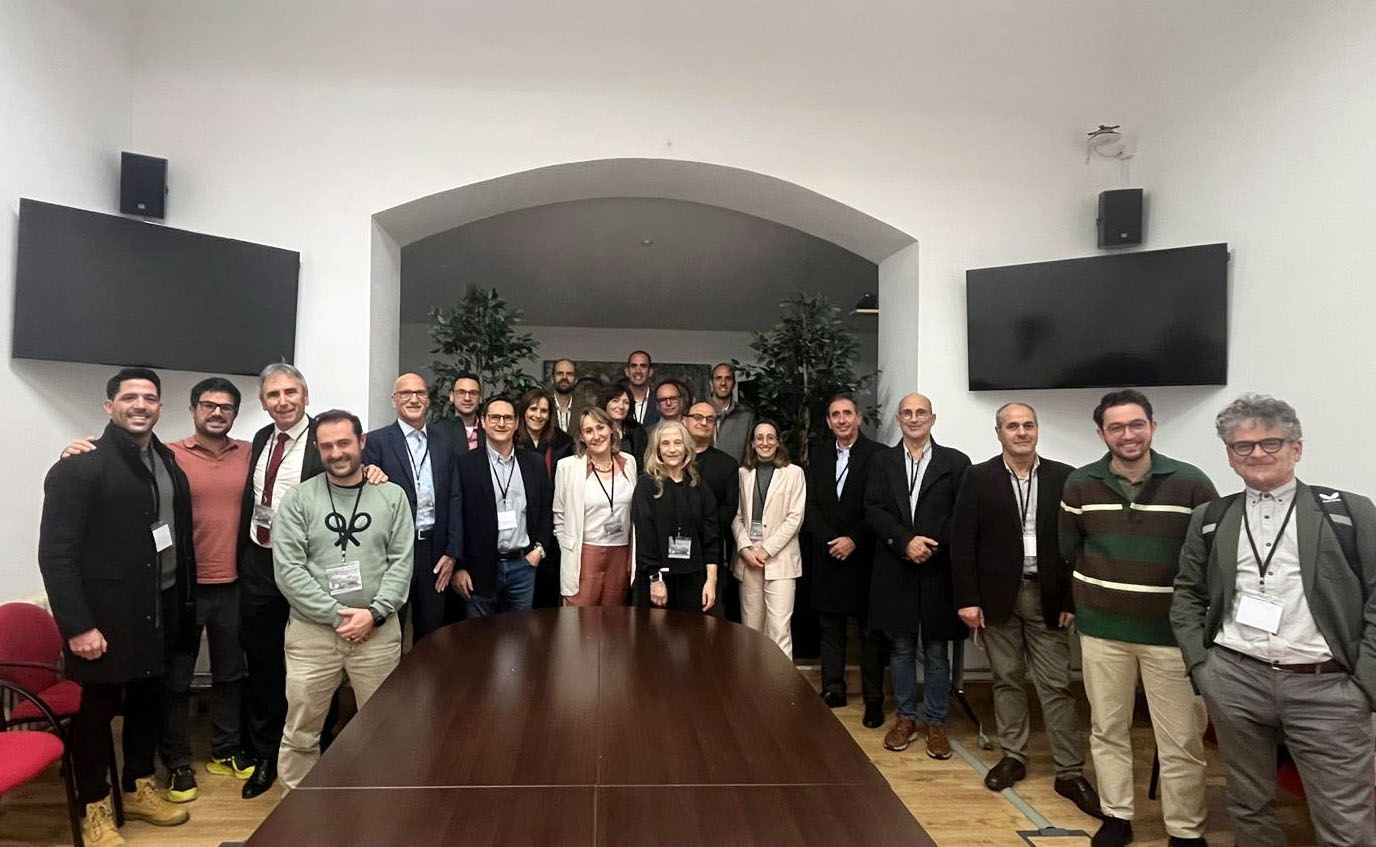 Un grupo numeroso de personas posando juntas para una foto al interior de una sala de reuniones. Hay una mesa de madera grande en primer plano, rodeada de sillas rojas. La mayoría de los presentes visten ropa formal, como trajes, sacos y credenciales colgadas al cuello, lo que sugiere que podrían ser participantes de una conferencia o reunión profesional. Al fondo de la sala hay dos plantas decorativas y dos pantallas planas en las paredes laterales. El ambiente es amistoso y todos los presentes están sonriendo.
