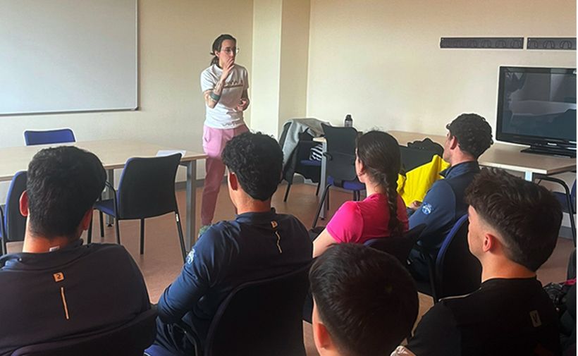 Una sala de aula con varias personas sentadas de espaldas, atendiendo a una persona de pie al frente. La persona que habla está cerca de una pizarra blanca grande en la pared izquierda; lleva camiseta clara y pantalón rosa, con el cabello recogido, y tiene una mano cerca de la boca mientras sostiene algo pequeño en la otra. Frente a ella hay mesas y sillas azules y grises.  En primer plano hay al menos seis personas sentadas en sillas, la mayoría con camisetas oscuras; una persona en el centro lleva una camiseta rosa y tiene el pelo largo recogido en una coleta. A la derecha se ve un televisor o monitor grande sobre una mesa; encima, en la pared, hay una franja oscura con dibujos o iconos repetidos. En una mesa cerca del frente hay una chaqueta y una botella.