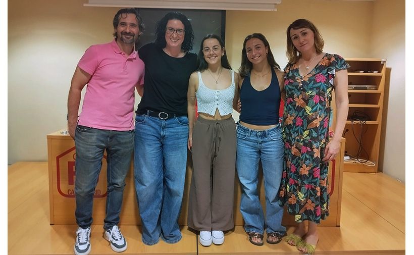 Cinco personas posando juntas en una sala. Hay tres mujeres jóvenes en el centro y dos adultos, un hombre a la izquierda y una mujer a la derecha. El hombre lleva una camiseta polo rosa y jeans, y sonríe. La mujer a su lado lleva gafas, una camiseta negra y pantalones vaqueros anchos. La joven en el centro viste un top blanco sin mangas y pantalones marrones anchos, y sonríe. A su lado, otra joven lleva un top negro y jeans claros. La mujer a la derecha lleva un vestido largo con estampado floral y sandalias verdes. Todos parecen relajados y contentos. El fondo muestra una mesa de madera clara y una estantería.