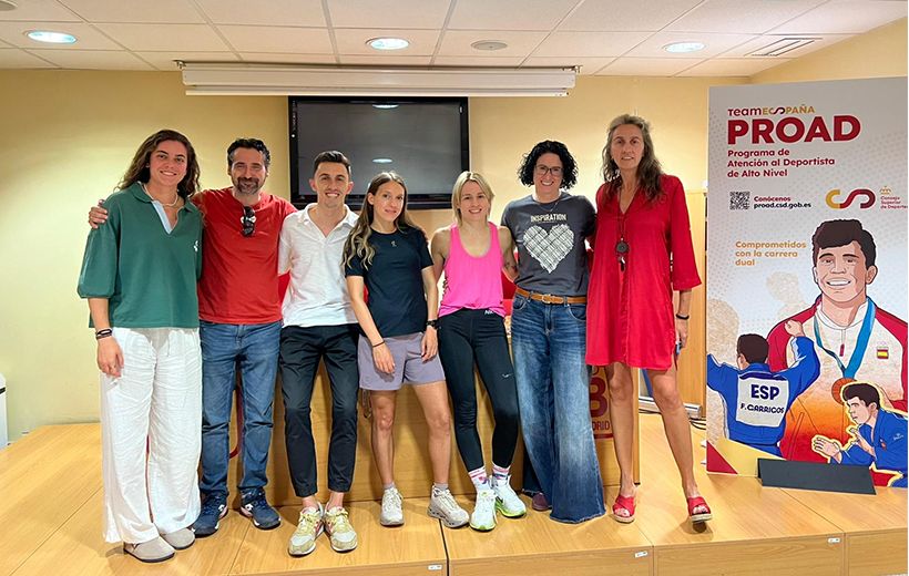 Grupo de siete personas posando sobre una tarima de madera en una sala interior. Están alineadas mirando hacia el frente, de cuerpo entero, sonriendo y con los brazos en algunos casos sobre los hombros de quienes tienen al lado.  De izquierda a derecha: Una mujer alta con piel clara, pelo castaño claro y ondulado hasta los hombros, lleva una blusa verde amplia con manga tres cuartos y pantalón ancho blanco, con zapatos beige. Un hombre de piel clara, pelo corto oscuro con algo de canas y barba, viste camiseta roja, vaqueros azules y zapatillas oscuras, con gafas de sol colgadas del cuello. Un hombre de piel clara y pelo corto castaño, lleva camisa blanca de manga larga remangada, pantalón oscuro ajustado y zapatillas claras con detalles verdes. Una mujer joven de piel clara y pelo largo castaño, lleva camiseta negra de deporte, falda corta gris deportiva y zapatillas deportivas claras. Otra mujer joven de piel clara y pelo rubio recogido, viste camiseta sin mangas rosa intensa, mallas negras ajustadas y zapatillas deportivas beige. Una mujer de piel clara con pelo rizado oscuro a la altura de la mandíbula lleva gafas, una camiseta gris con un diseño claro en el centro, vaqueros azules amplios y zapatillas deportivas oscuras. A la derecha, una mujer muy alta de piel clara y pelo castaño claro algo ondulado viste un vestido corto rojo de manga larga tres cuartos y sandalias rojas con tacón.  Al fondo, la pared es color crema y hay un televisor negro apagado colgado en el centro. El techo es blanco con focos empotrados redondos. A la derecha de la imagen se ve un gran cartel vertical del programa PROAD, con el texto “team ESPAÑA PROAD Programa de Atención al Deportista de Alto Nivel” y otros textos más pequeños, junto a ilustraciones de deportistas con ropa de España, predominando los colores rojo, blanco, azul y amarillo.