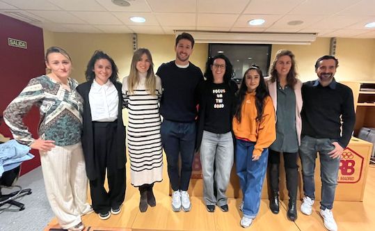 La imagen muestra a ocho personas de pie, posando para una foto en un interior. Están alineadas en una fila y parecen estar en un ambiente de oficina o sala de conferencias. La persona en el centro lleva una camiseta negra con el texto "FOCUS ON THE GOOD". A la izquierda de la imagen, hay un letrero verde que dice "SALIDA". Las personas están sonriendo y parecen estar en un ambiente amigable. Al fondo, hay una pared de color beige y un podio con el logo de BB Madrid.