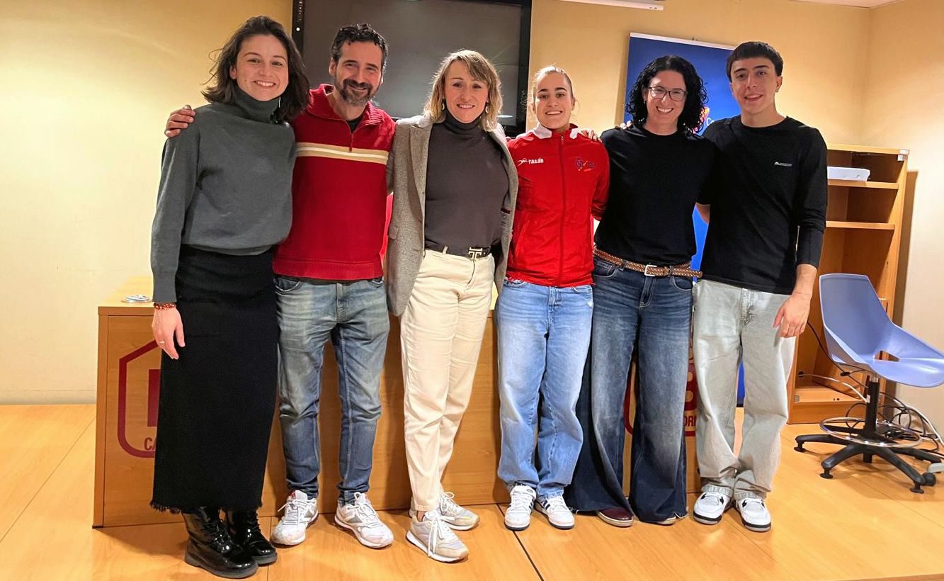 Grupo de seis personas de pie, abrazándose y sonriendo a la cámara en una sala de aspecto profesional o académico. Al fondo hay una mesa de madera, una pantalla de televisión apagada y un estante con una silla vacía a la derecha. La iluminación es cálida y el ambiente parece relajado y amistoso. Todas las personas visten ropa informal; destacan los colores neutros, rojo y negro en sus atuendos.