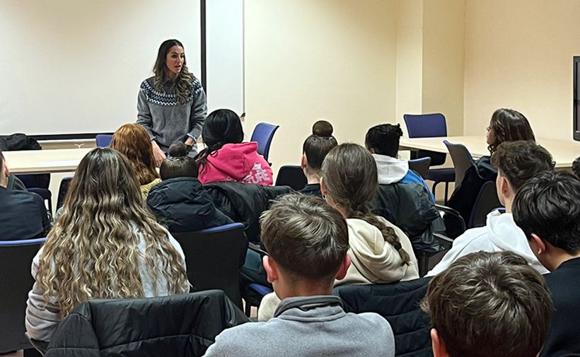 Aula de clase con una mujer de cabello largo y suéter gris de pie frente a una pizarra blanca, hablando a un grupo de estudiantes sentados. La mujer es la gimnasta Carolina Rodríguez. Los estudiantes están de espaldas, algunos con chaquetas puestas y varios con el cabello largo. El ambiente es de aprendizaje en una sala con paredes claras y sillas azules. Hay una mesa al frente junto a Carolina Rodríguez y otra más hacia el fondo a la derecha. La atención de los alumnos está centrada en ella mientras comparte su experiencia.